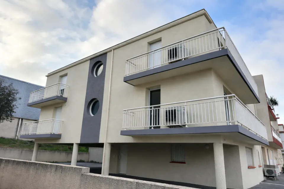 Façade d’un immeuble de trois niveaux : enduit crème, balcons à garde-corps blancs et bande verticale grise percée de deux hublots