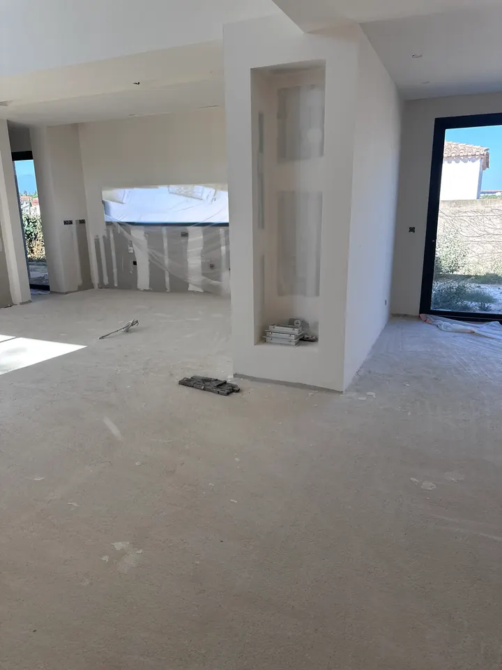 Séjour aux murs et plafonds blancs uniformisés après ratissage, niche encore en plaque de plâtre brute, film plastique sur la fenêtre de la cuisine et sol brut