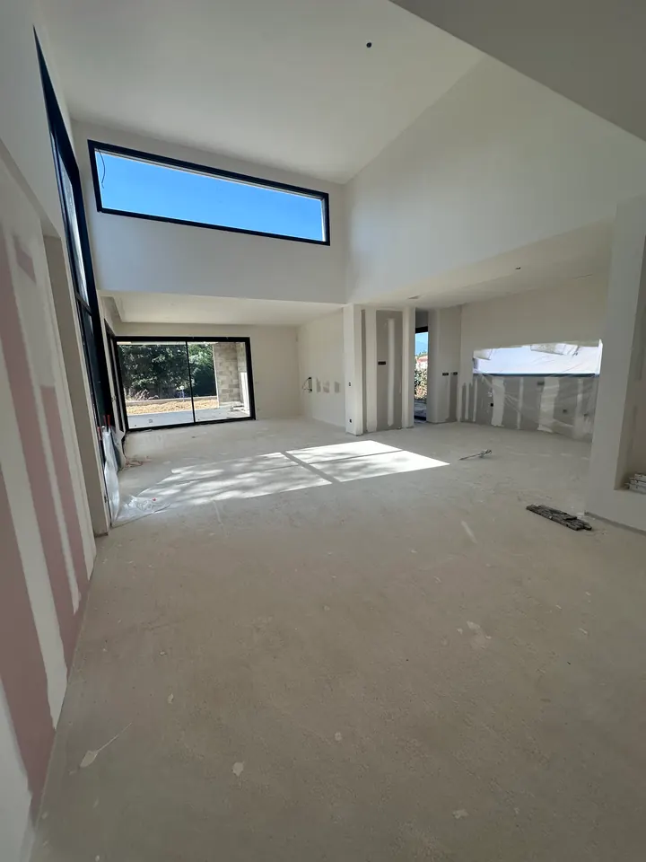 Grand séjour sous haut plafond aux murs blancs uniformisés après ratissage, haute fenêtre horizontale, sol brut et parois encore protégées d'un film en fond de pièce