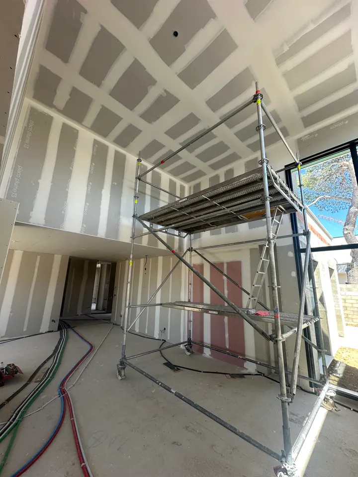 Grand volume sous haut plafond avant travaux : murs et plafond en plaques de plâtre aux bandes de joints visibles, échafaudage roulant et grande baie vitrée