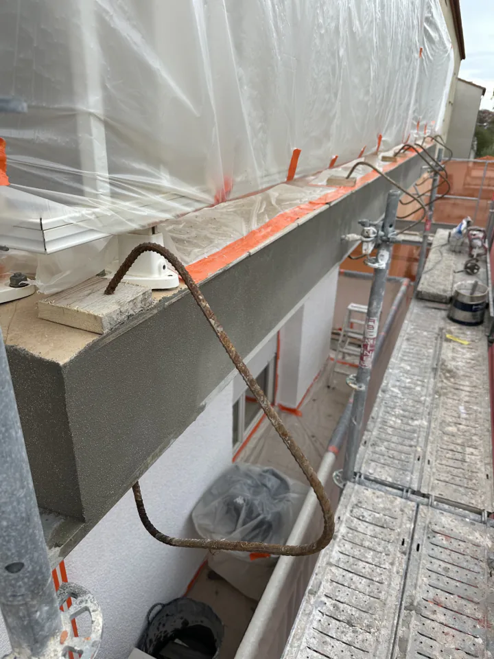 Rive de balcon enduite de gris avec une armature métallique rouillée qui dépasse, protections plastiques et échafaudage autour