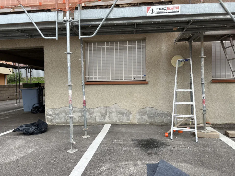 Partie basse d'une façade avec fenêtre à barreaux et blocs apparents, sous un échafaudage