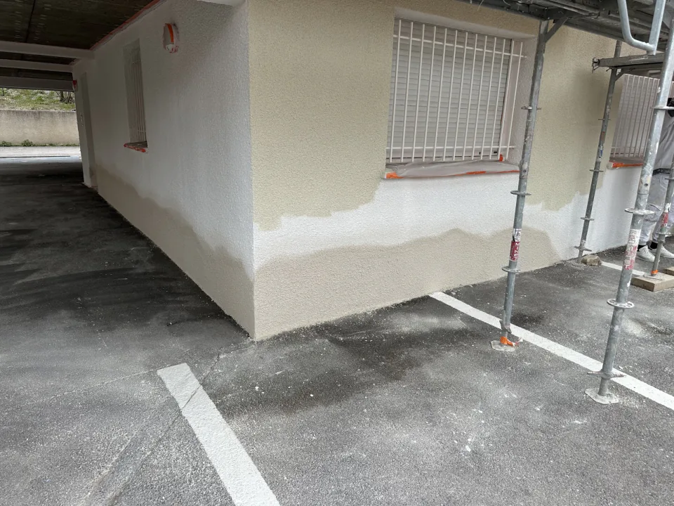Angle de façade en cours de peinture : partie basse beige et partie haute blanche, fenêtre à barreaux et échafaudage